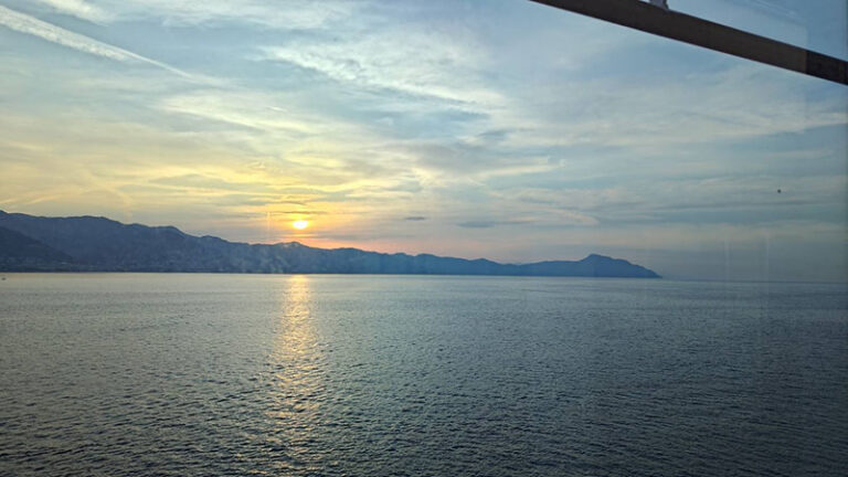 「クルーズ船上にて、地中海の朝陽を初めて迎える」 暁がほのかに開き、地中海の天蓋にはなお夜の余韻が漂っている。 雲は初めの一筋の陽光にそっと金の粉を染められ、海面は静かな鏡のように凪ぎ、 ただ遠く、旭日が柔らかな黄金の小径を引き、山影の懐へとまっすぐ延びている。